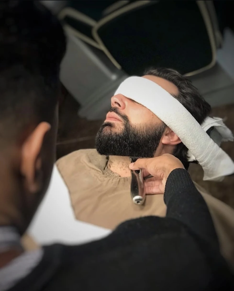 Jonathan trabalhando na barba de um cliente com toalha sobre os olhos